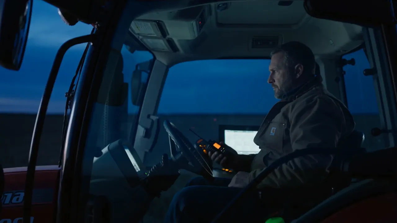 A farmer keying an Icom two-way radio in a tractor cab on a Manitoba field at dusk